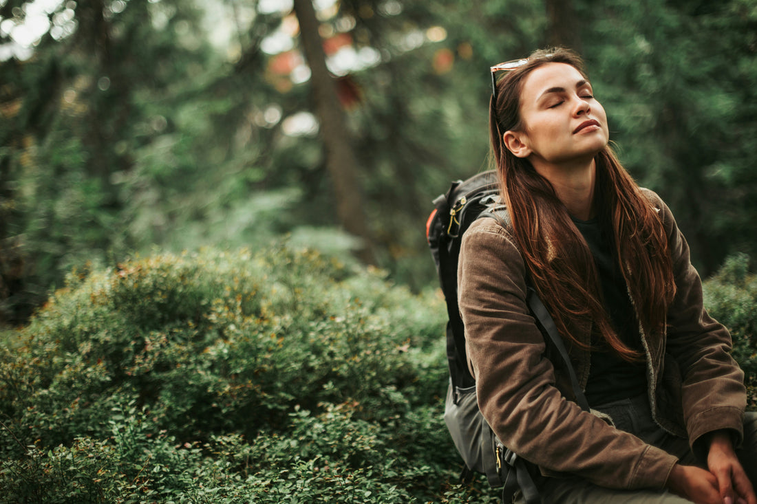 A woman resting outdoors in a forest, eyes closed, wearing a brown corduroy jacket with a backpack